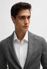 Gray wool blend blazer with notched lapels, single chest pocket, and subtle texture, worn over a white button-up shirt.