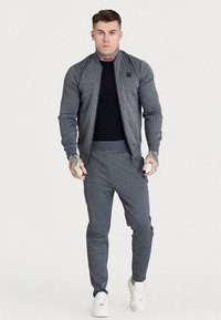 Casaco bomber cinzento texturizado com design canelado, usado sobre uma camisa preta; combinado com calças cinzentas a condizer e ténis brancos.