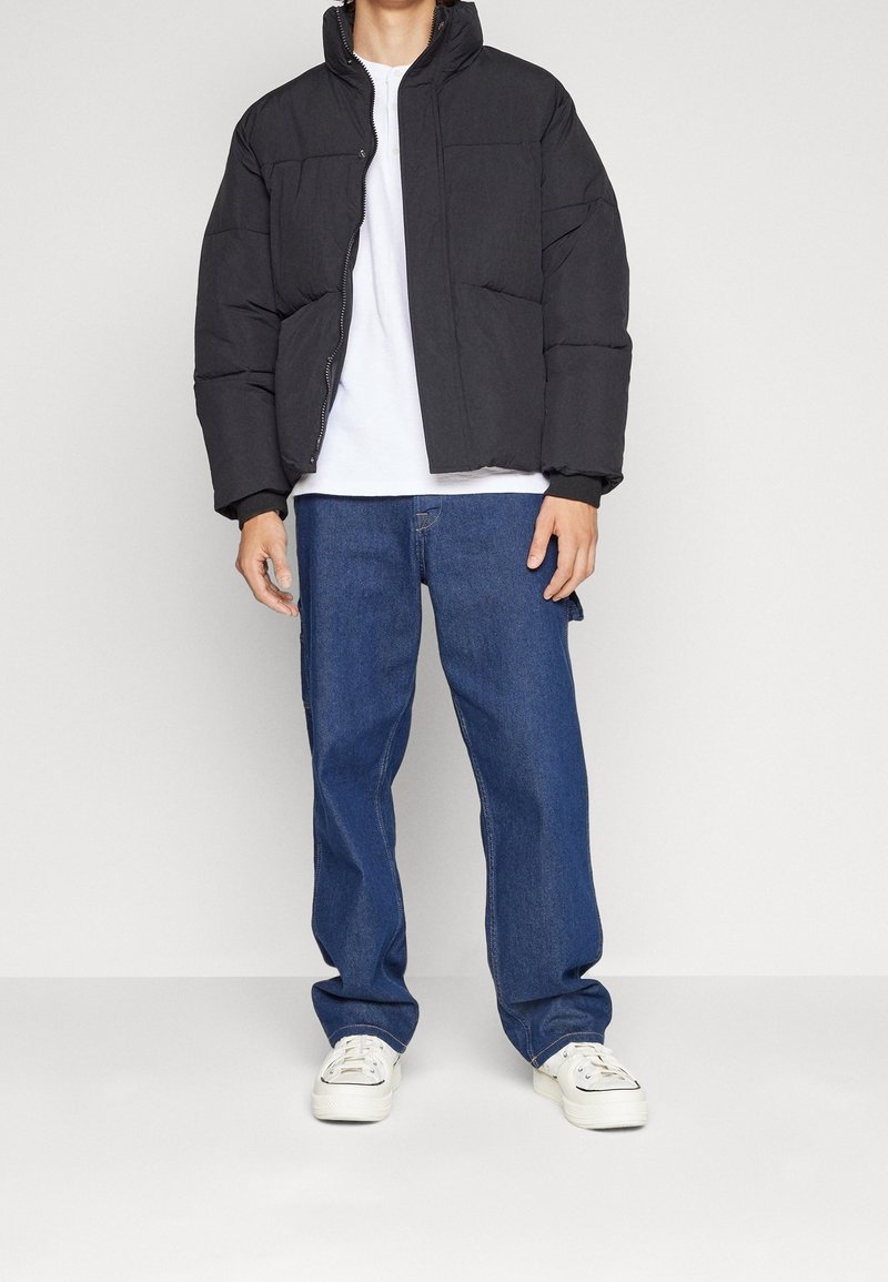 Personne portant une doudoune noire par-dessus une chemise blanche avec un jean bleu ample et des baskets blanches, debout devant un fond clair uni.