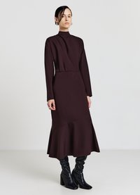 Langärmliges, hochgeschlossenes Kleid in dunklem Burgunderrot, mit einem ausgestellten Saum und einer taillierten Passform, hergestellt aus glattem Stoff. Kombiniert mit schwarzen kniehohen Stiefeln.