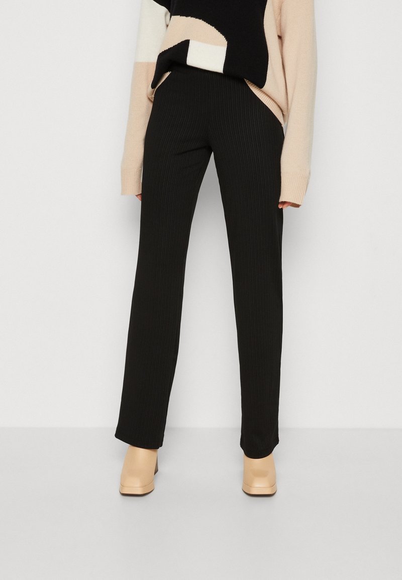Schwarze Rippengamaschen mit geradem Beinschnitt. Aus strukturiertem Stoff, kombiniert mit einem beige-schwarz gemusterten Pullover und braunen Absätzen.