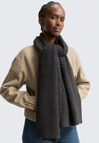 Dunkelgrauer Schal aus strukturiertem Stoff, um den Hals gewickelt. Das Model trägt einen beigen Pullover und blaue Jeans.