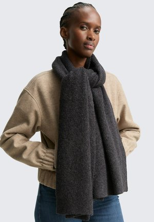 Écharpe gris foncé en tissu texturé, enroulée autour du cou. La mannequin porte un pull beige et un jean bleu.