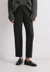 Persoon in zwarte op maat gemaakte broek, zwarte glanzende loafers en een grijze grofgebreide trui tegen een effen muur.