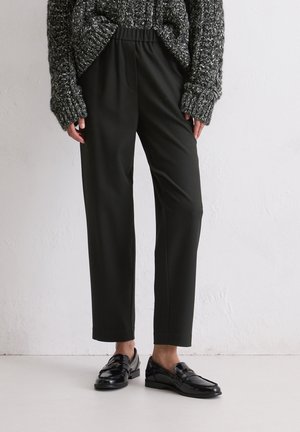 Persoon in zwarte op maat gemaakte broek, zwarte glanzende loafers en een grijze grofgebreide trui tegen een effen muur.