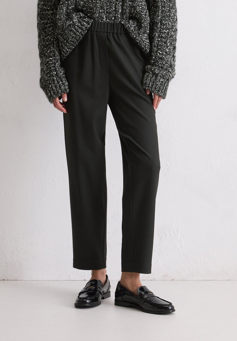 Persoon in zwarte op maat gemaakte broek, zwarte glanzende loafers en een grijze grofgebreide trui tegen een effen muur.