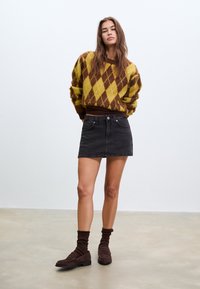 Jeune femme portant un pull à losanges marron et jaune, une mini-jupe en jean noir, des chaussettes marron et des mocassins marron, debout sur un sol uni.