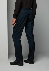 Marineblauwe broek met een getailleerde pasvorm, een gladde textuur, twee achterzakken en een subtiele glans. Gecombineerd met zwarte nette schoenen.