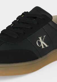 Gros plan sur une basket en cuir noir avec des lacets noirs, une semelle en gomme et le logo blanc "cK" sur le côté.
