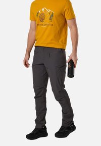 Άνδρας που φοράει μουσταρδί κίτρινο outdoor graphic t-shirt και γκρι παντελόνι πεζοπορίας με φερμουάρ, κρατώντας ένα μαύρο μπουκάλι νερού στο δεξί του χέρι.