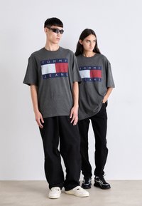 Camisetas gráficas grises con un logo rectangular en azul marino, blanco y rojo. Combinadas con pantalones de pierna ancha negros y zapatillas de contraste.