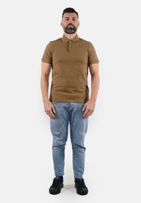 Polo shirt marrone con colletto, abbinato a jeans chiari strappati e sneakers nere. Il modello è in piedi di fronte con le mani ai fianchi.