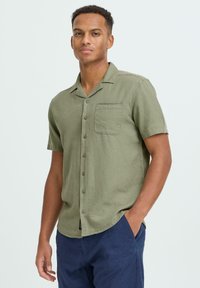 Chemise en lin vert olive à manches courtes avec une poche poitrine, fermeture à boutons et coupe décontractée, associée à un pantalon bleu marine.