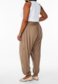 Brązowe, luźne spodnie haremowe z szerokim pasem, elastycznymi mankietami i gładką teksturą, w połączeniu z białą bezrękawnikową bluzką i płaskimi butami.