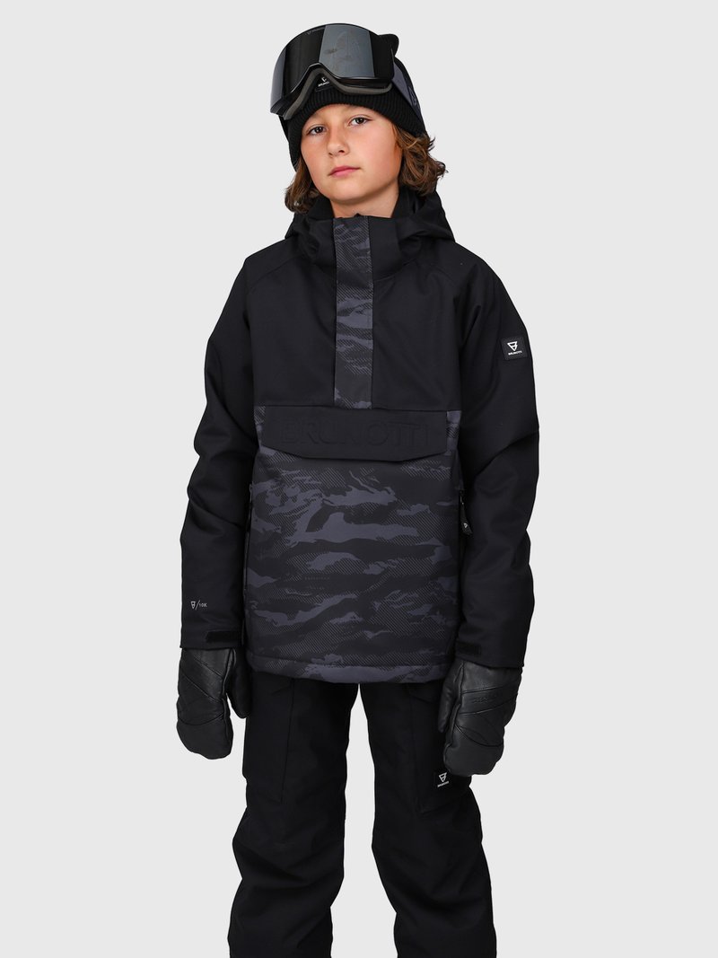 Niño vestido con una chaqueta de esquí negra con patrón de camuflaje, pantalones de esquí negros, guantes negros, un gorro negro y gafas de esquí sobre el casco, frente a un fondo gris.