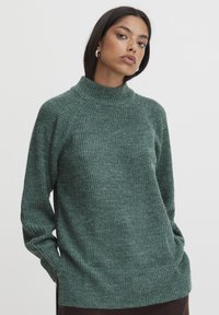 Grüner, gestrickter Pullover mit hohem Kragen, Raglanärmeln, strukturiertem Stoff und Seitenschlitzen am Saum.