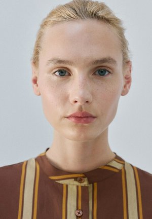 Jonge vrouw met blond haar in een staart, blauwe ogen, neutrale uitdrukking, draagt een bruin shirt met verticale beige en gele strepen.