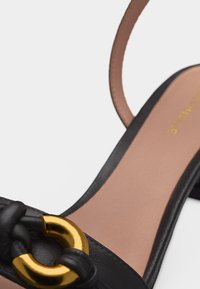 Μαύρο δερμάτινο σανδάλι με χρυσή κυκλική αγκράφα στο μπροστινό λουράκι και ταμπά εσωτερική σόλα με ορατό ράψιμο.