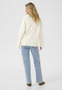 Gebreide crème trui met een scalloped zoom en een ontspannen pasvorm, gecombineerd met lichtblauwe jeans met een diagonale zakontwerp en bruine schoenen.