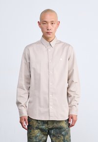 Camisa botón de color beige claro con cuello, mangas largas y un pequeño logo en el pecho. Acabado de tejido suave, combinada con pantalones de patrón camuflaje.
