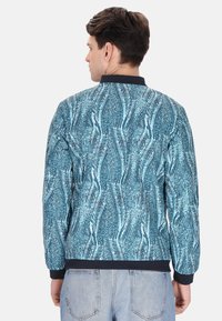 Giacca da uomo con un motivo blu texturizzato, caratterizzata da un colletto e polsini neri a coste. Il design presenta linee fluide simili a onde.