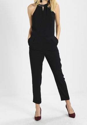 Femme portant une combinaison noire sans manches à col halter avec poches et des talons hauts bordeaux, debout devant un fond uni.