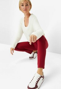White ribbed long-sleeve top, red fitted pants, and white sneakers with burgundy accents. The model is squatting with a hand on her leg.