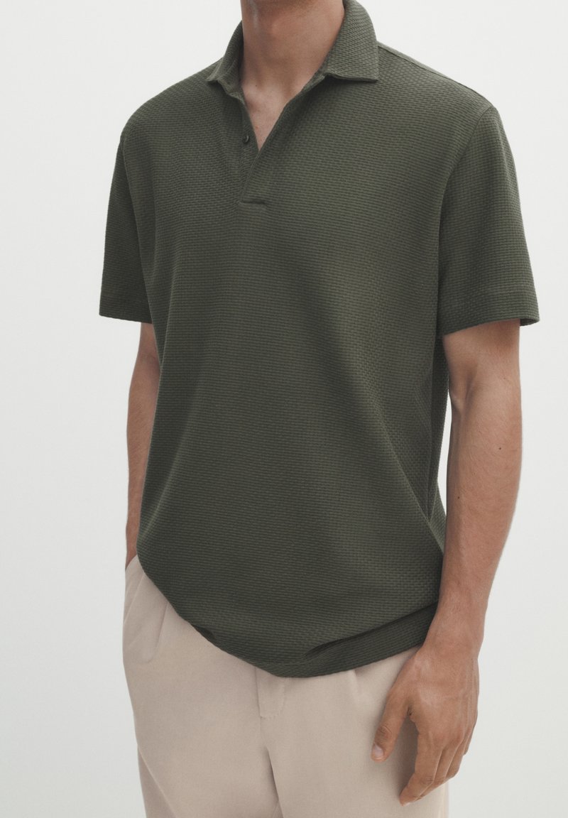 Polo shirt vert foncé avec un tissu texturé, manches courtes, patte de boutonnage à trois boutons et col classique. Associé à un pantalon beige clair.