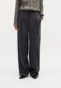 Weite Hosen aus dunkelgrauem Tweed mit Vorderfalten; kombiniert mit einem karierten Button-Up-Shirt in Grau und Gelb. Schwarze spitze Schuhe runden den Look ab.