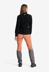 Kvinna som står vänd bort, iklädd en svart långärmad topp och tvåfärgade bruna och grå byxor med svarta skor.