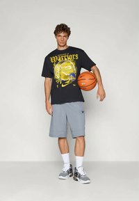 Juodas Nike marškinėlis su geltona "Warriors" spauda ir grafika. Pilkos sportinės šortai, baltos kojinės ir pilki/juodi sportbačiai. Laiko krepšinį.