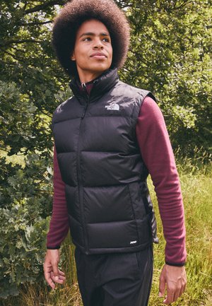 Veste rembourrée noire avec un col haut, fermeture éclair, et un logo en haut à gauche. Portée par-dessus une chemise à manches longues bordeaux.