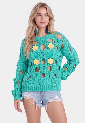 Donna con lunghi capelli biondi che indossa grandi orecchini di piume, un maglione a rete aperta turchese con dettagli floreali gialli e pantaloncini di jeans strappati.