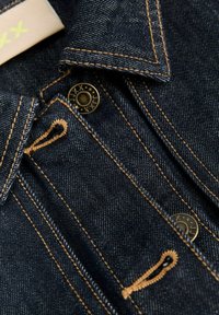 Tumesinist teksajope lähedalt, kus on metallist nupud tähte ja "JXX" kujundusega ning nupusilmade ümber kontrastne oranž õmblus.