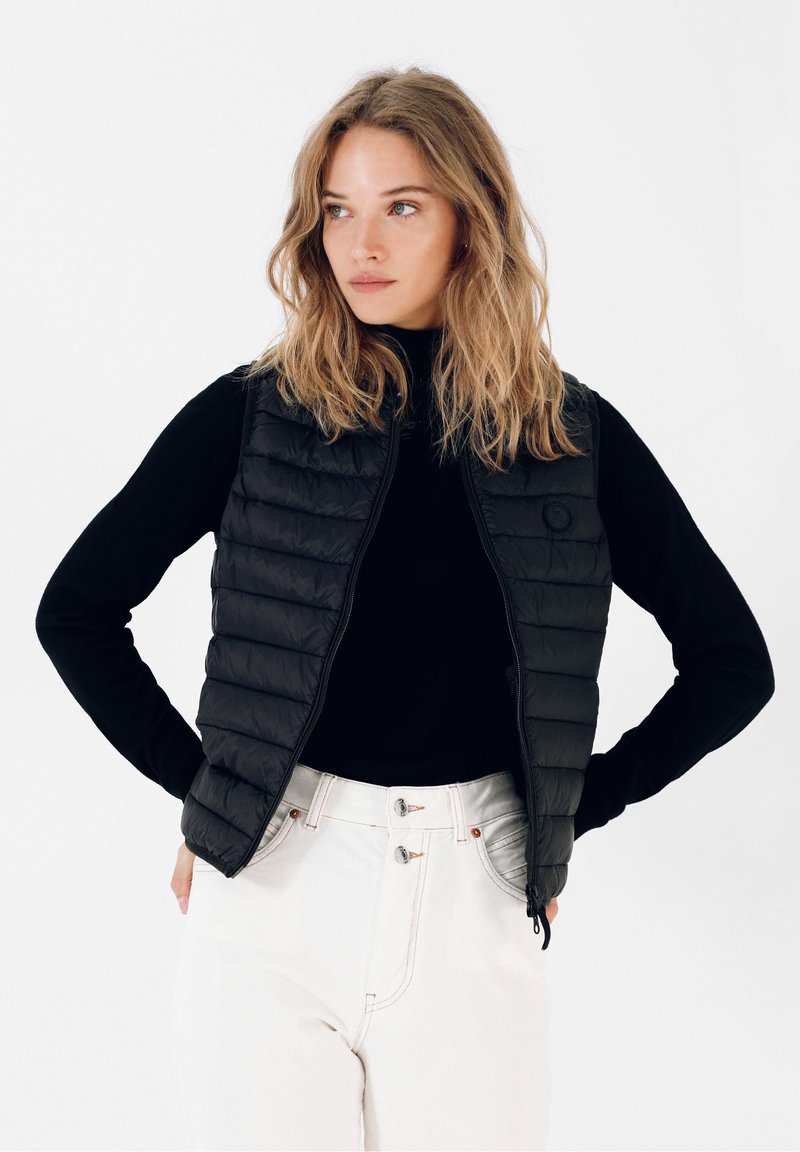 Sort puffer vest med quiltet mønster, lynlås lukning og rundt logo. Båret over en sort turtleneck og parret med hvide bukser.