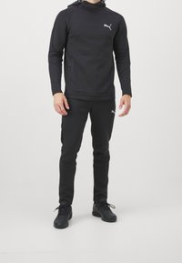 Sweat à capuche et pantalon de sport noirs en matière douce, avec une poche kangourou et des accents de logo, assortis à des chaussures de sport noires.