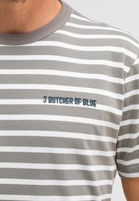 Grau und weiß gestreiftes T-Shirt mit Rundhalsausschnitt und kurzen Ärmeln; auf der Brust mit "J BUTCHER OF BLUE" in Dunkelblau bedruckt.