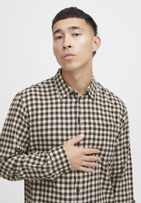 Camisa de franela a cuadros marrón y blanco con botones en el frente, cuello y un bolsillo en el pecho. Textura suave con mangas largas.