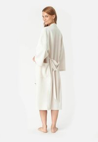 Helle beigefarbene Robe mit Bindegürtel, weiten Ärmeln und seitlichen Taschen. Glatte Textur, knielanges Design. Rückansicht des Models.