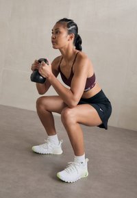 Sort kettlebell, tilpasset maroon sports-bh, sorte shorts, hvide sneakers med neongrøn sål. Indendørs fitnesscenter med neutrale toner.
