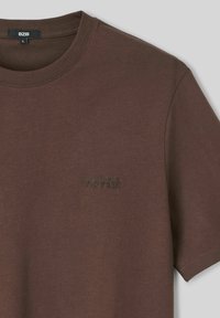 T-shirt en coton marron avec un col ras du cou et des manches courtes. Doté d'un texte brodé "no risk" sur la poitrine gauche. Texture lisse.