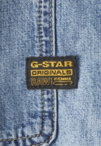 Etykieta dżinsowa G-Star Originals z czarnym tłem i złotym napisem. Materiał ma jasnoniebieski kolor z widocznymi detalami przeszyć.