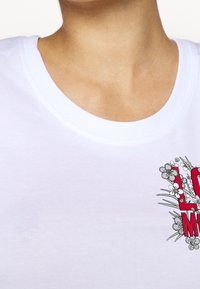Vit t-shirt med kort ärm som har rund halsringning och ett tryckt mönster med texten "LOVE" i rött, omgiven av blommönster.