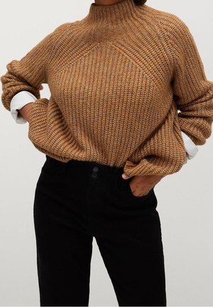 Personne portant un pull en maille marron texturé avec manches retroussées sur une chemise blanche, associé à un pantalon noir taille haute, mains dans les poches.