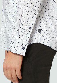 Witte blouse met lange mouwen, voorzien van een zwart abstract patroon, met knoopmanchetten en een klein logo-label nabij de zoom. Zwarte broek is gedeeltelijk zichtbaar.