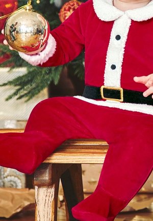 Bébé vêtu d'un costume de Père Noël rouge tenant une décoration de Noël dorée, assis sur un banc en bois près d'un arbre décoré.