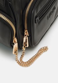 Bolso de mano de cuero negro con dos cremalleras doradas, acento de cadena dorada y un logo en relieve en el costado. Textura suave con forma estructurada.