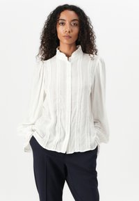 Vit blus med texturerade ränder och delikata spetsdetaljer. Har en hög krage och puffade ärmar, knapplist framtill och avslappnad passform.