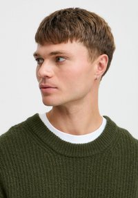 Dunkelgrüner Strickpullover mit gerippter Textur und weitem Rundhalsausschnitt, über einem weißen T-Shirt getragen. Kleine Hängerchenohrringe sichtbar.