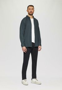 Dunkelgrüne Hemdbluse über einem weißen T-Shirt, kombiniert mit schwarzen Hosen und hellen Turnschuhen. Das Modell steht vor einem neutralen Hintergrund.